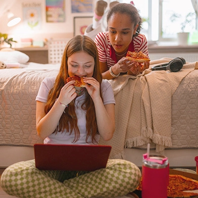 Two smiling teen girls eating pizza and looking at a laptop computer.