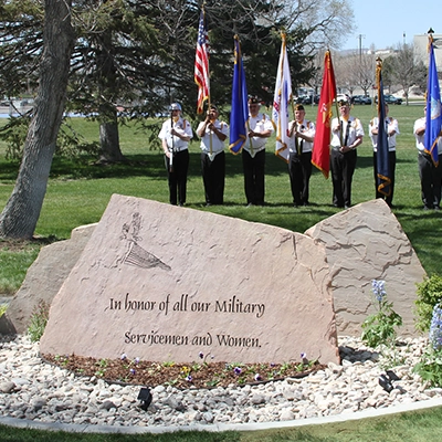 Stone monument with Military Appreciation Garden engraved on it with a background of a VFW Color Guard.