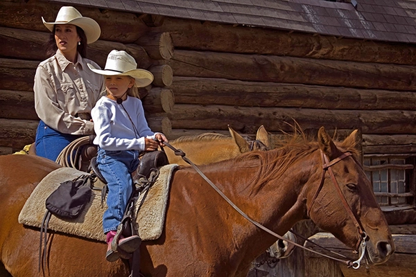 Mother and daughter each on a horse. Behind them is the wall a rustic ranch building.