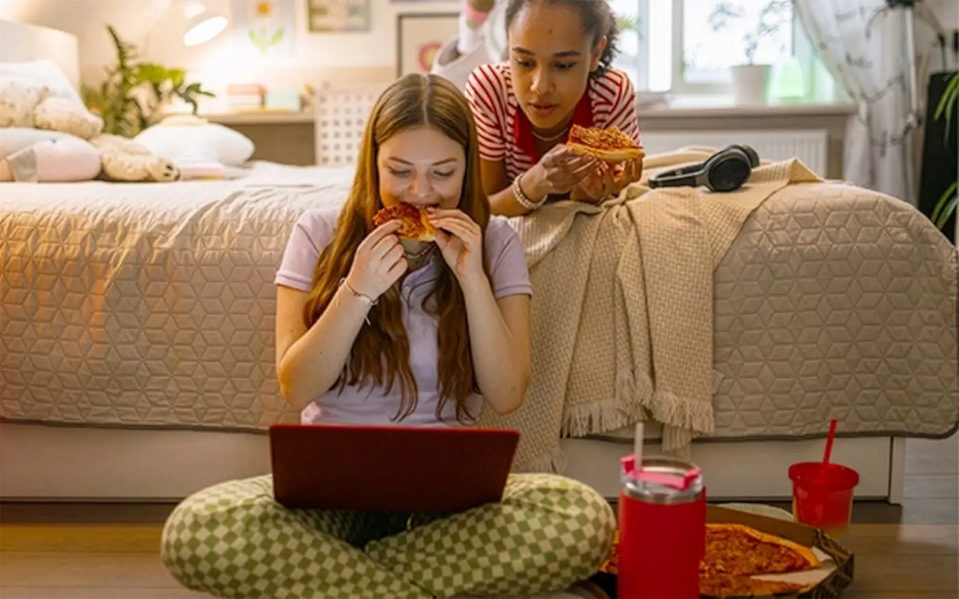 Two girls eating pizza while looking at a laptop screen.