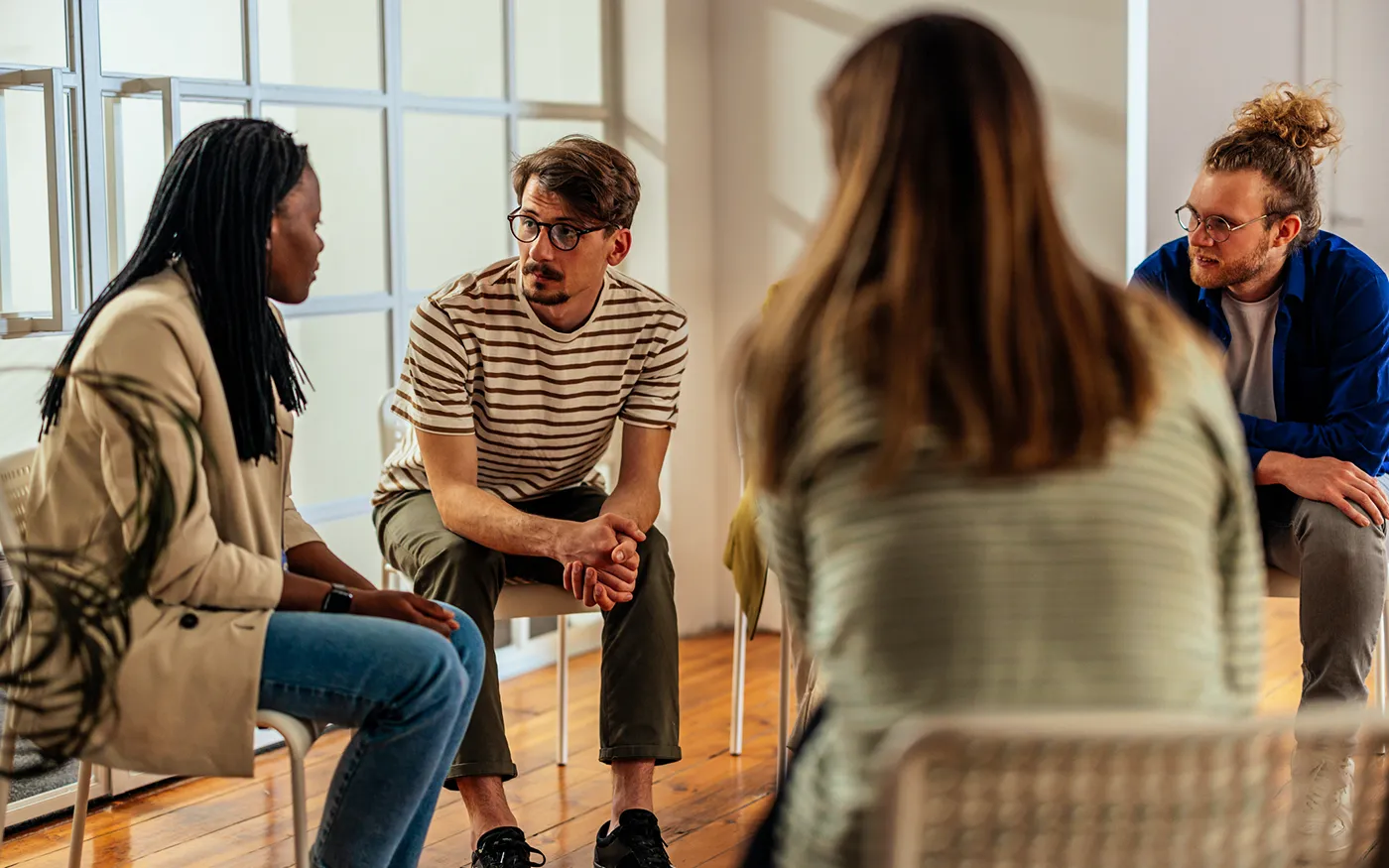 A group of people sit in a circle in an open, airy room, talking with each other.